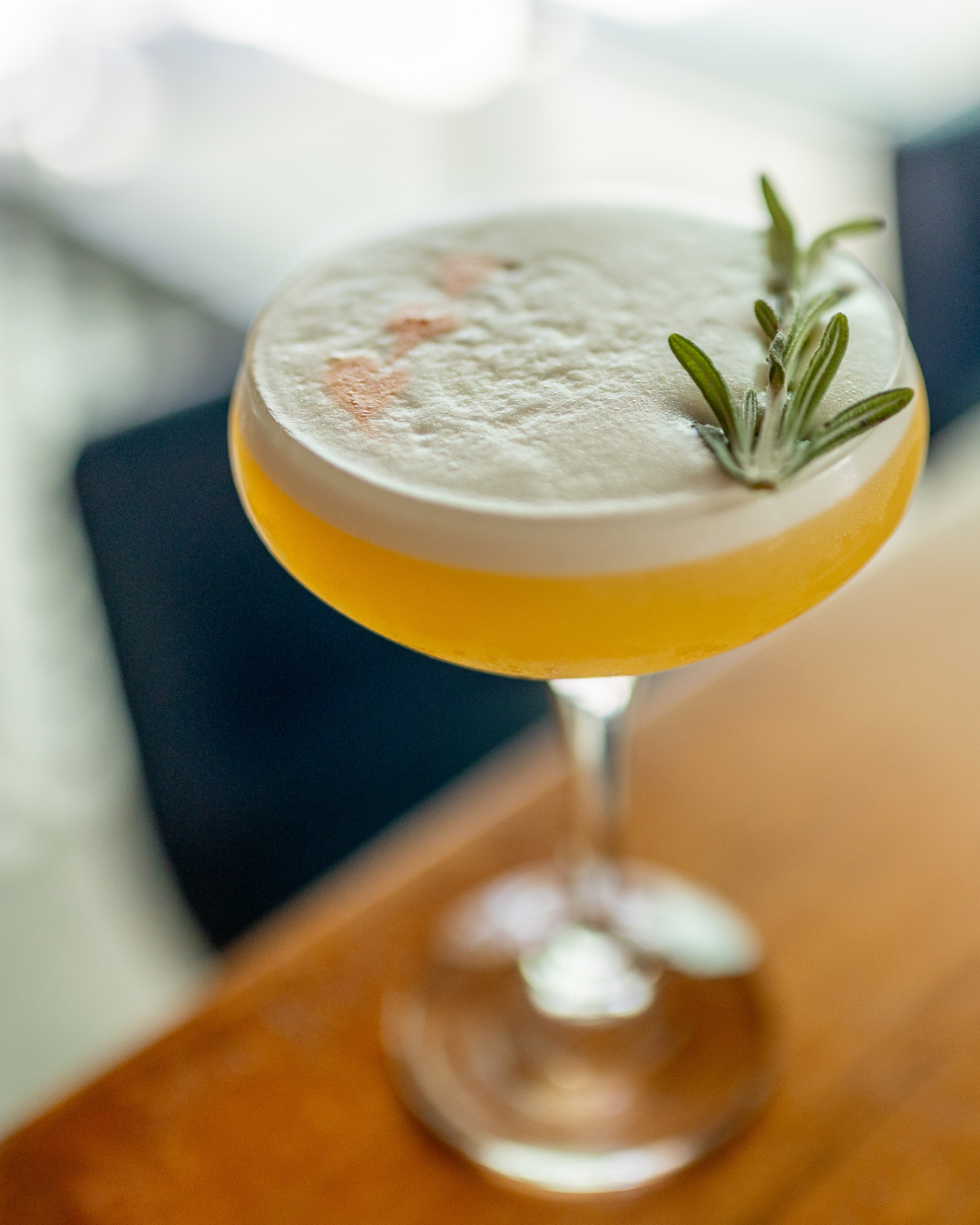 Craft cocktail with yellow citrus hue and herb garnish in a coupe glass.