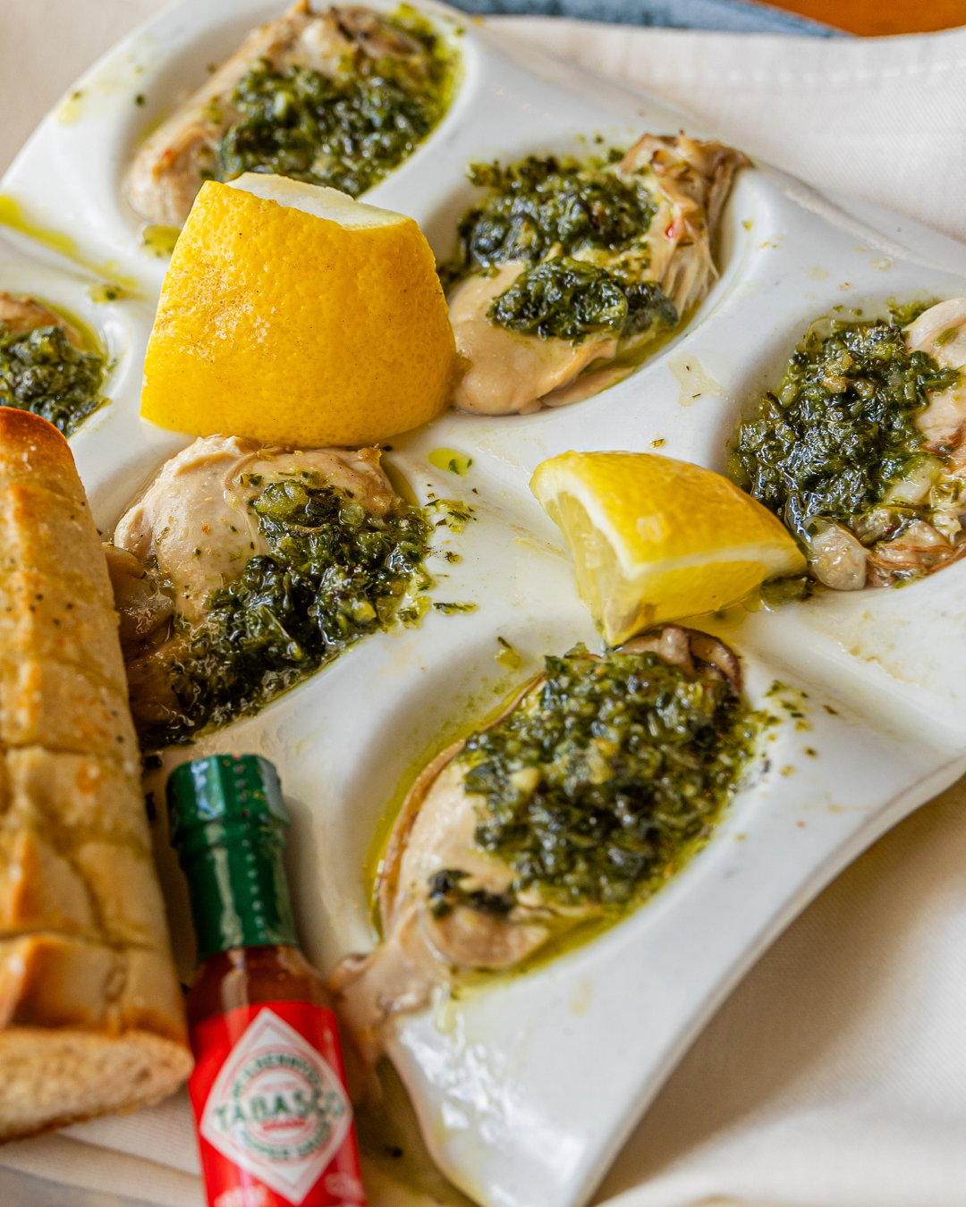 Fresh oysters topped with herbs and lemon wedges served on a white platter with a bottle of hot sauce.
