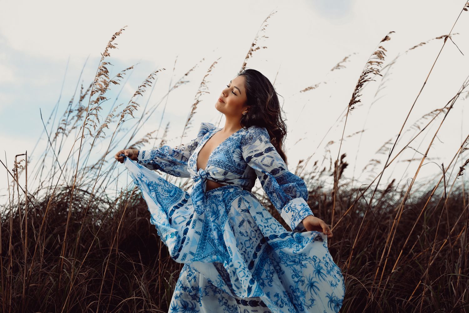 Person in flowing blue dress poses gracefully against tall grass and cloudy sky during outdoor photoshoot.