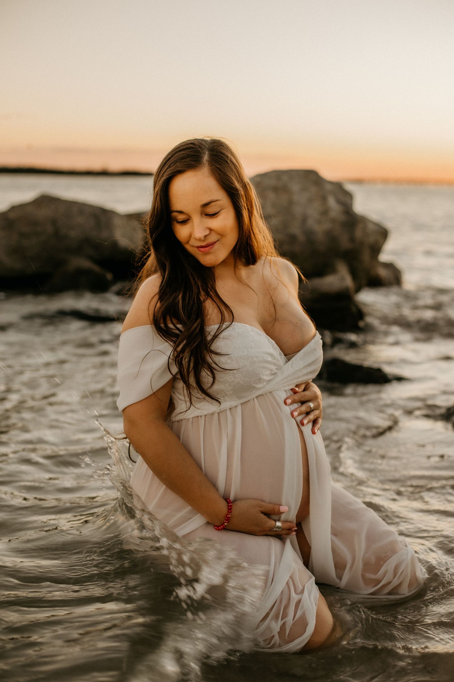 A flowing white dress maternity photo captured in shallow ocean water at sunset.