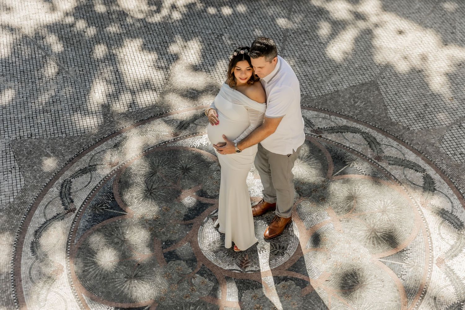 Two people embrace on a sun-dappled walkway, casting gentle shadows through trees above.