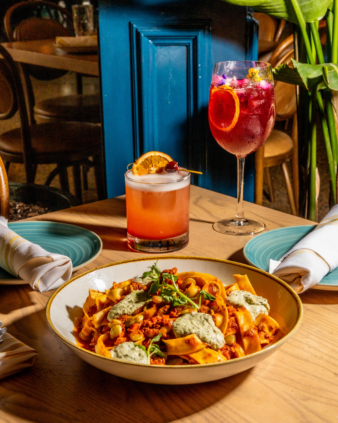 Pasta dish with red sauce and cocktails served on turquoise plates against blue wall.