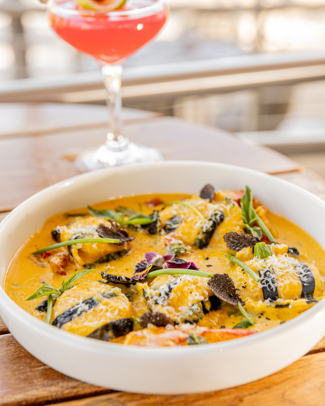 Creamy curry dish with black rolls and fresh herbs in a white bowl beside a pink cocktail.