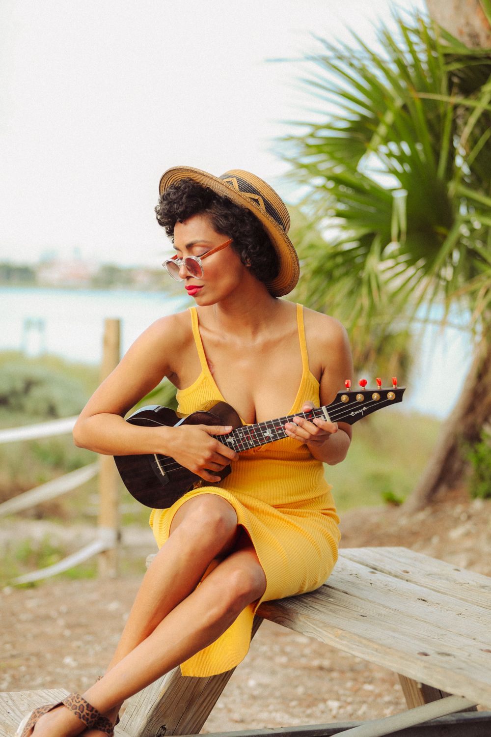 A person in a yellow sundress plays ukulele while sitting on a wooden beam at a tropical beach setting.