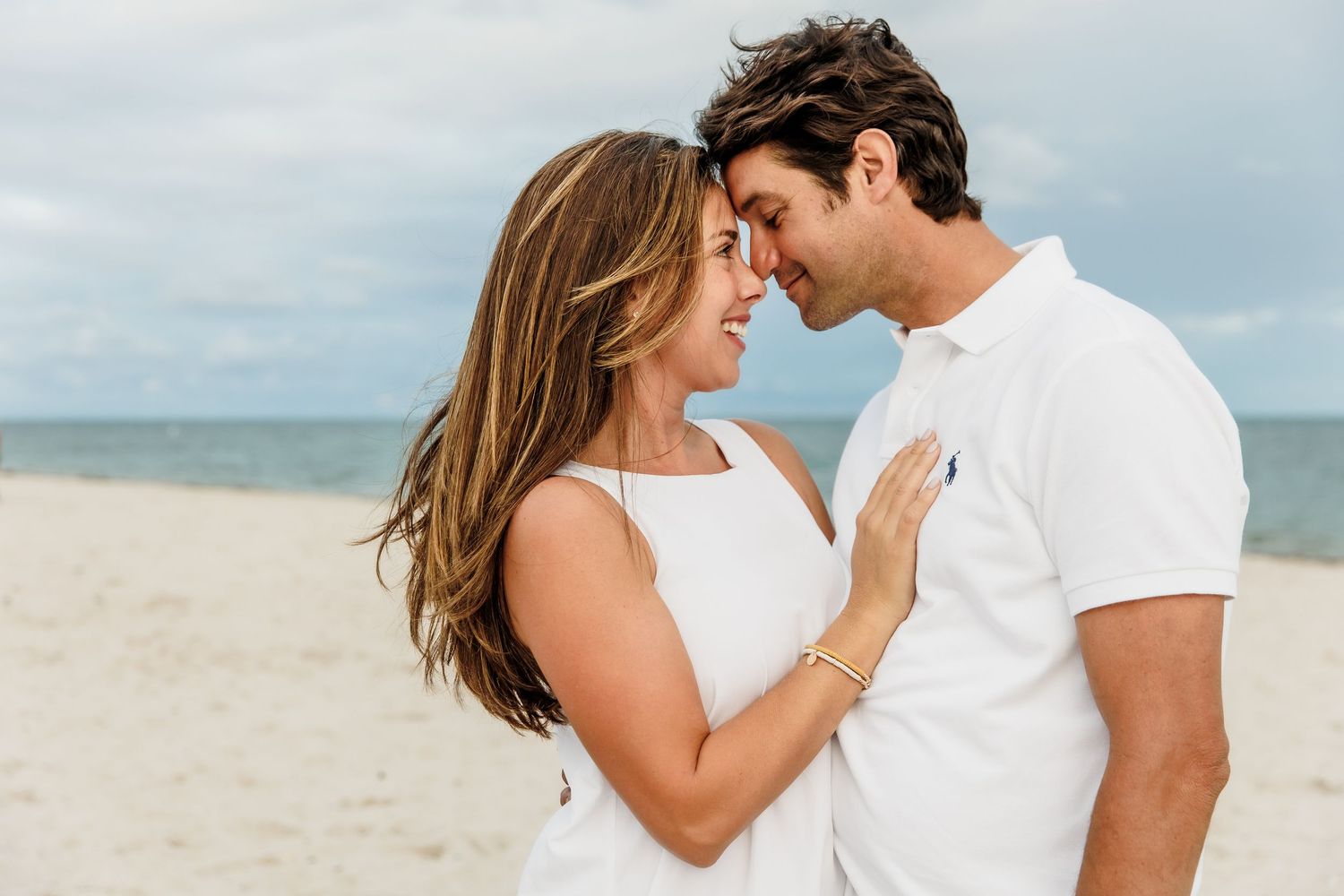 A romantic couple in white clothing embraces on a sunny beach with palm trees in the background.