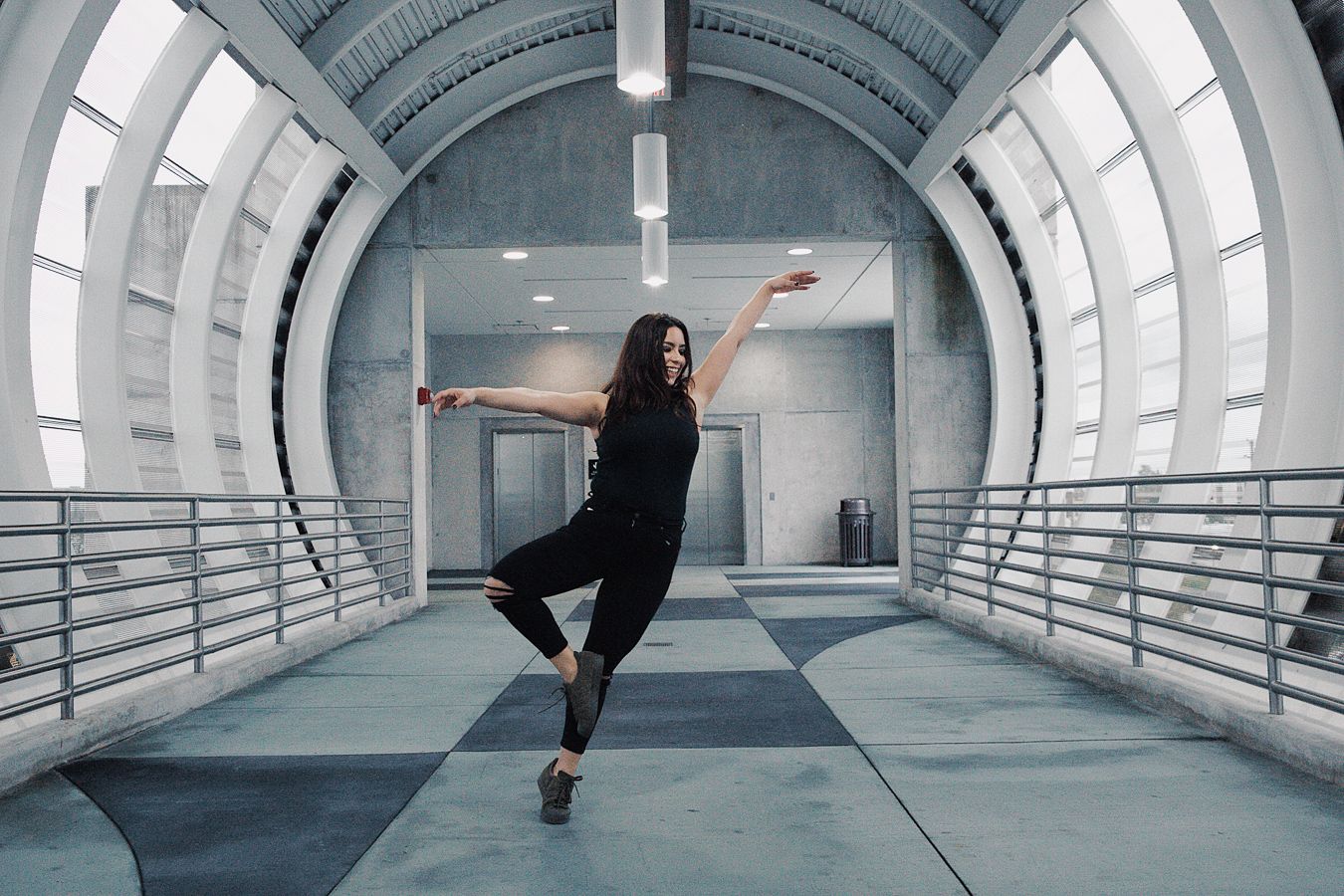 A dancer in black performs a graceful pose inside a modern tunnel with curved white walls and metal railings.