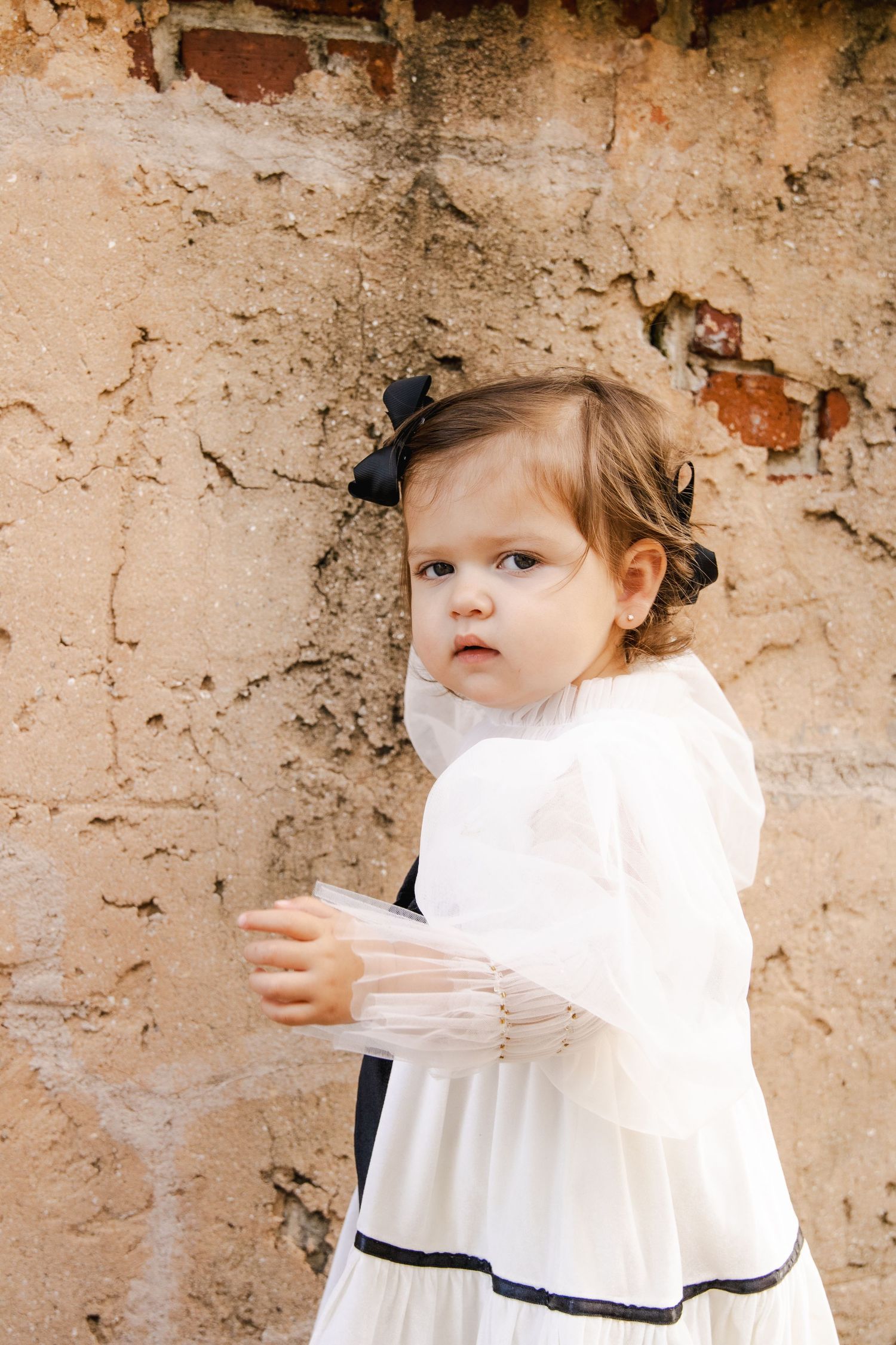 Child in white vintage dress stands against textured stone wall in artistic portrait series.