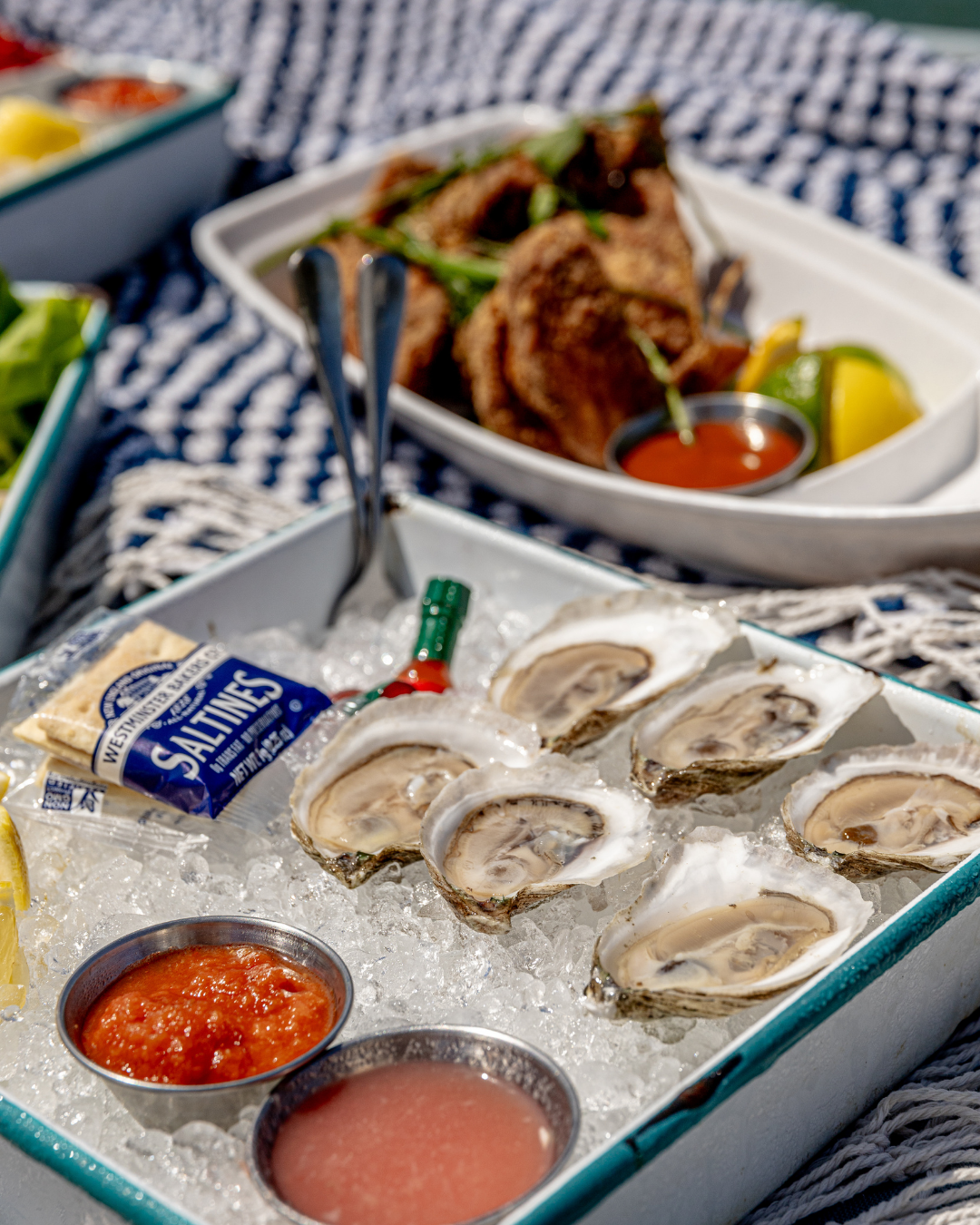Fresh oysters on ice with cocktail sauce and lemon wedges served on blue striped cloth.
