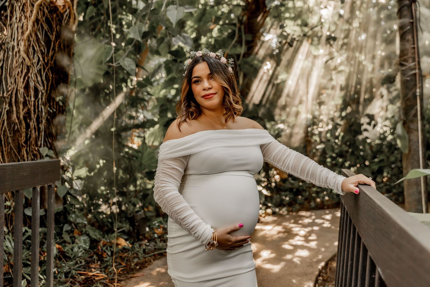 Expectant mother wearing an off-shoulder white dress poses in dappled sunlight on a wooden deck surrounded by trees.