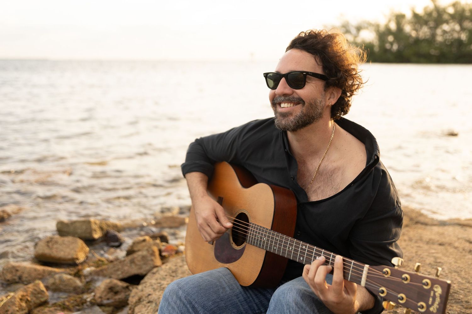 Musician in sunglasses plays acoustic guitar while sitting on rocky beach at sunset.