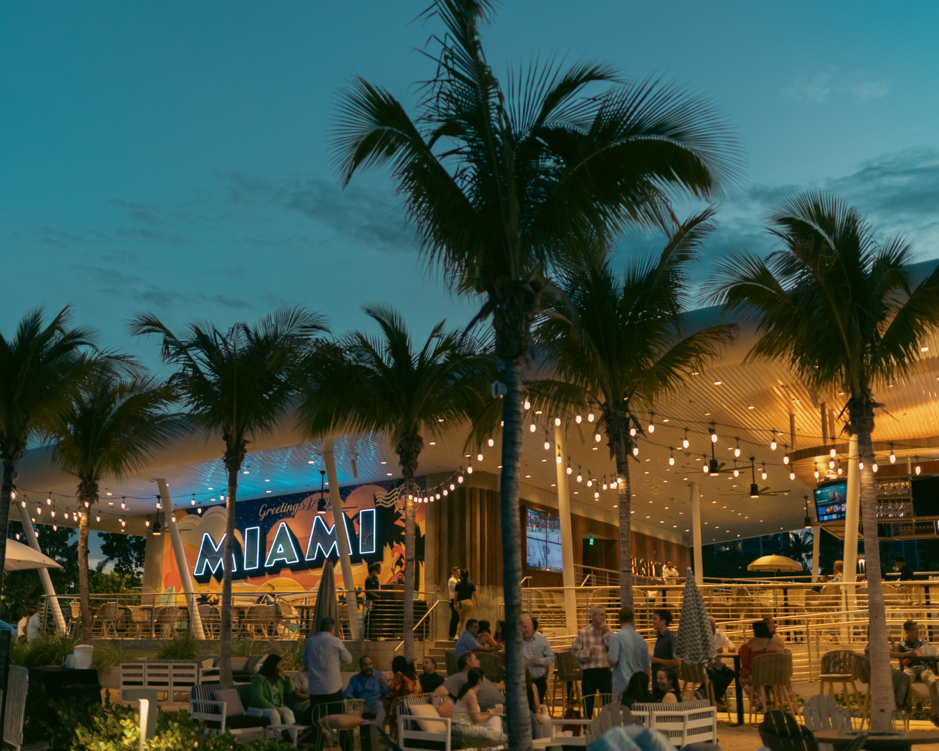 String lights illuminate a beachfront tiki bar at dusk with palm trees and a glowing sunset sky in the background.