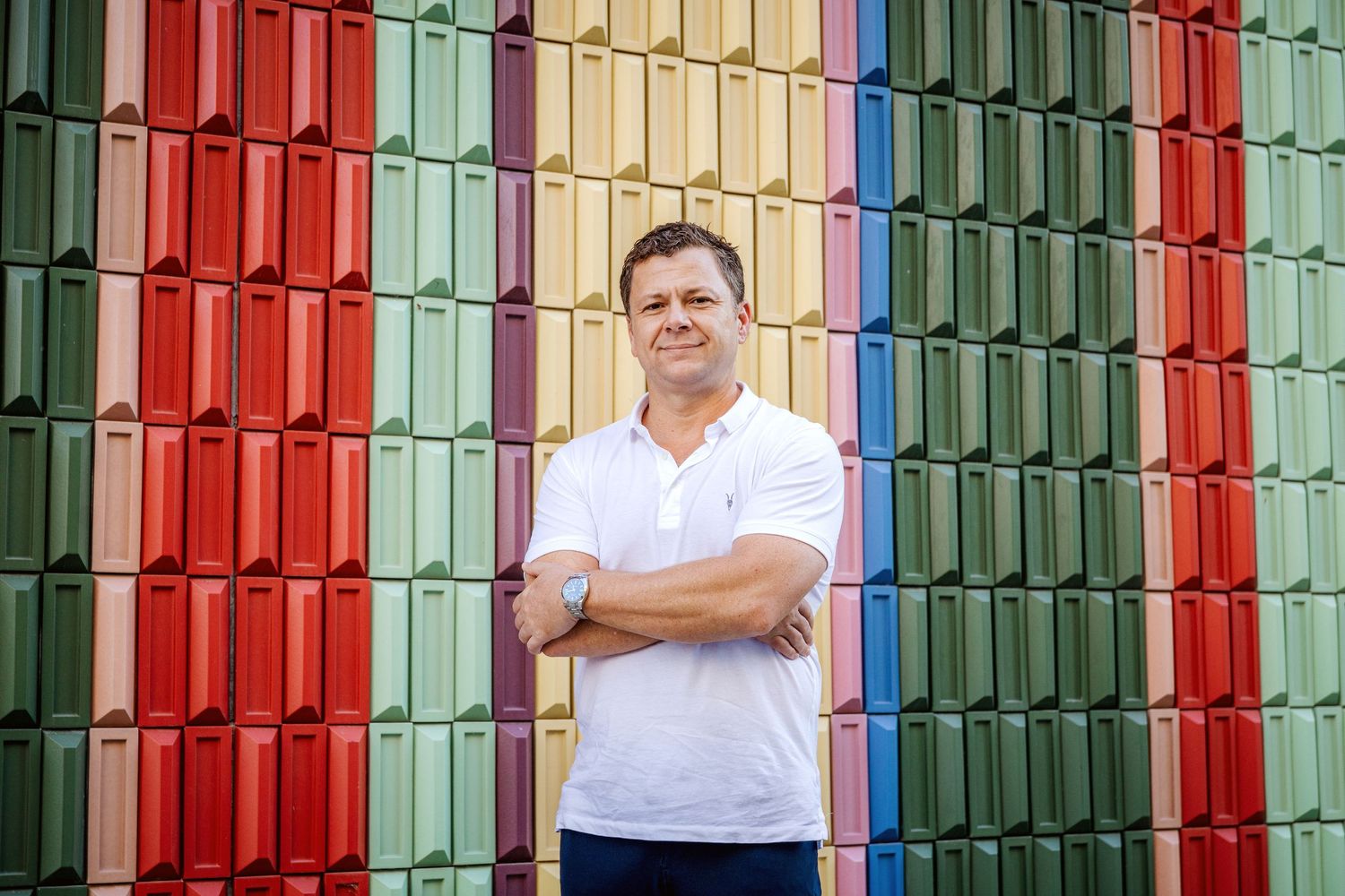 A figure stands in front of a vibrant wall decorated with colorful vertical slats in red, green, and beige tones.