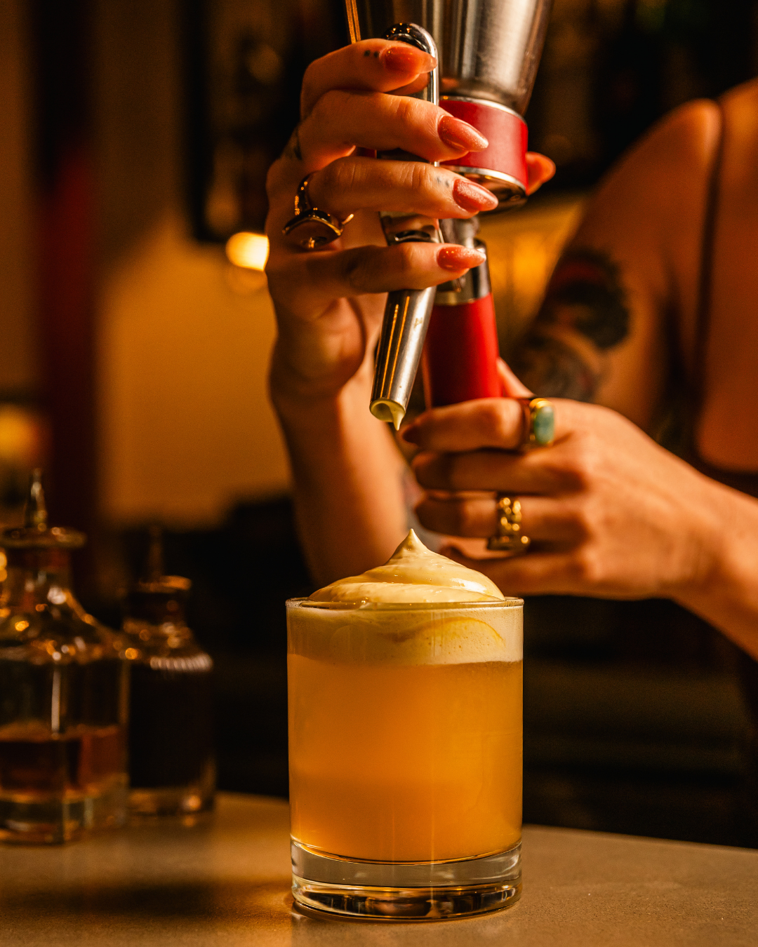 Hands topping a craft cocktail with foam using professional bartending tools in a cozy bar setting.