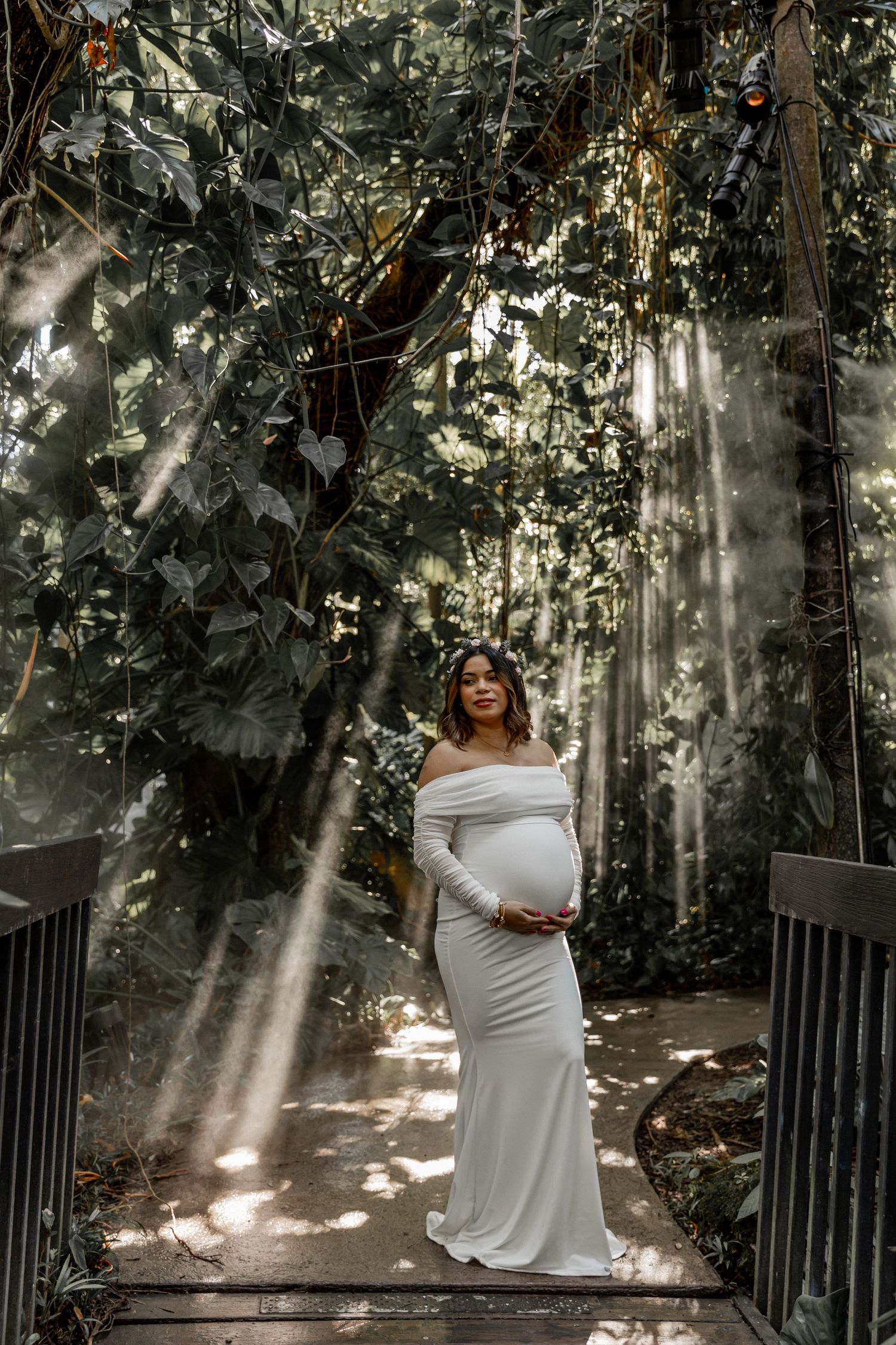 A serene maternity photoshoot in a garden setting with sunbeams filtering through trees and foliage.