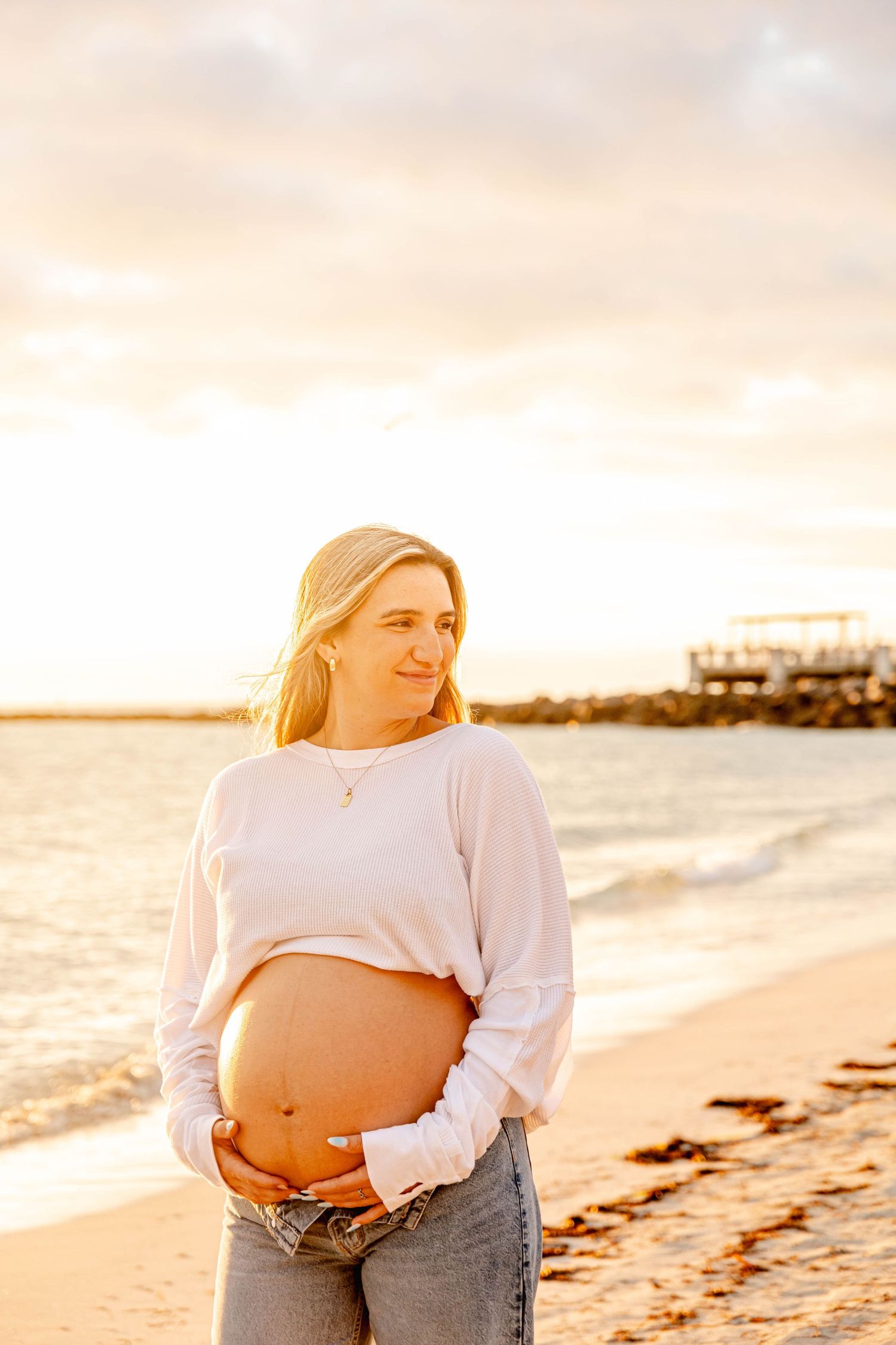 A beachside maternity portrait during golden hour with a pier in the background.