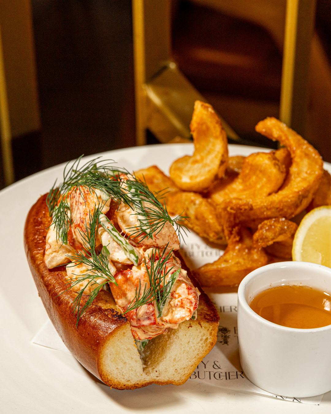 Lobster roll sandwich served with crispy potato wedges and dipping sauce on a white plate.