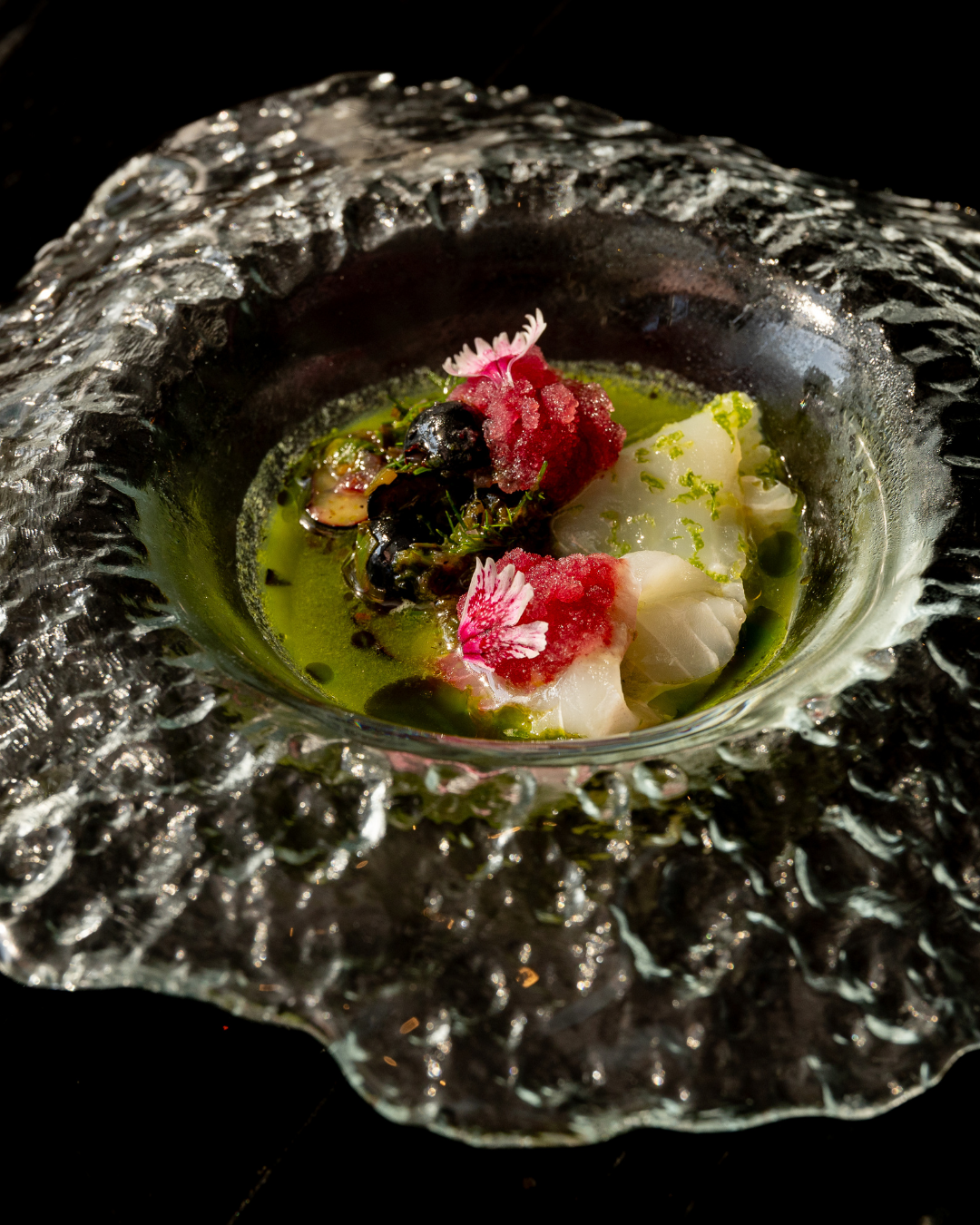 Artistic plating of seafood in green broth served in a textured glass bowl.