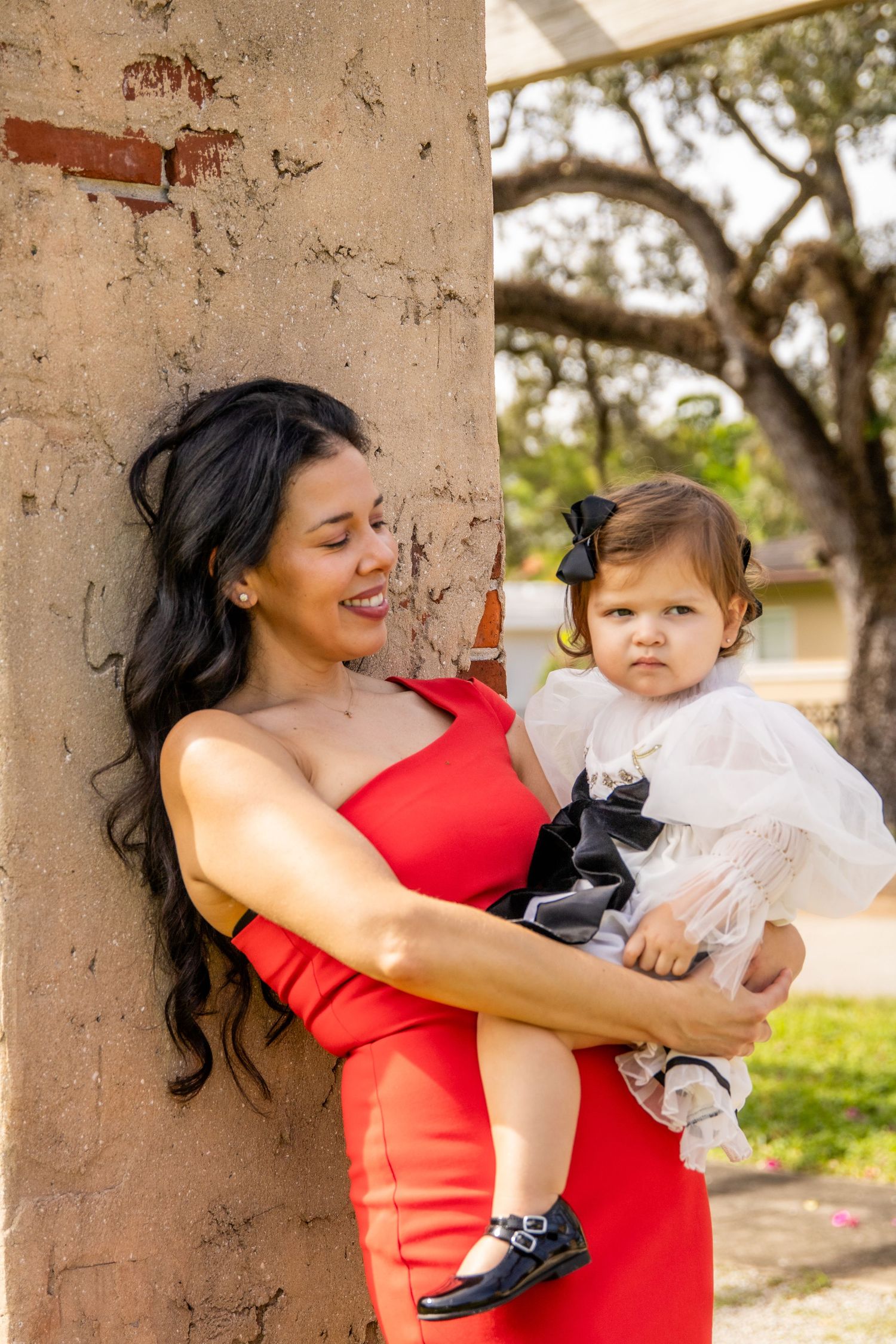 A woman in a red dress holds a baby in a white dress outdoors against a pillar.