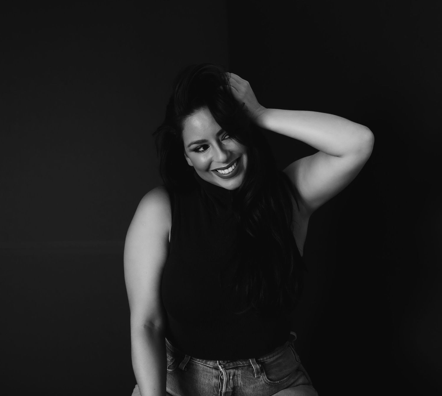 Black and white portrait of a woman in a sleeveless top and jeans smiling while running her hand through dark hair.