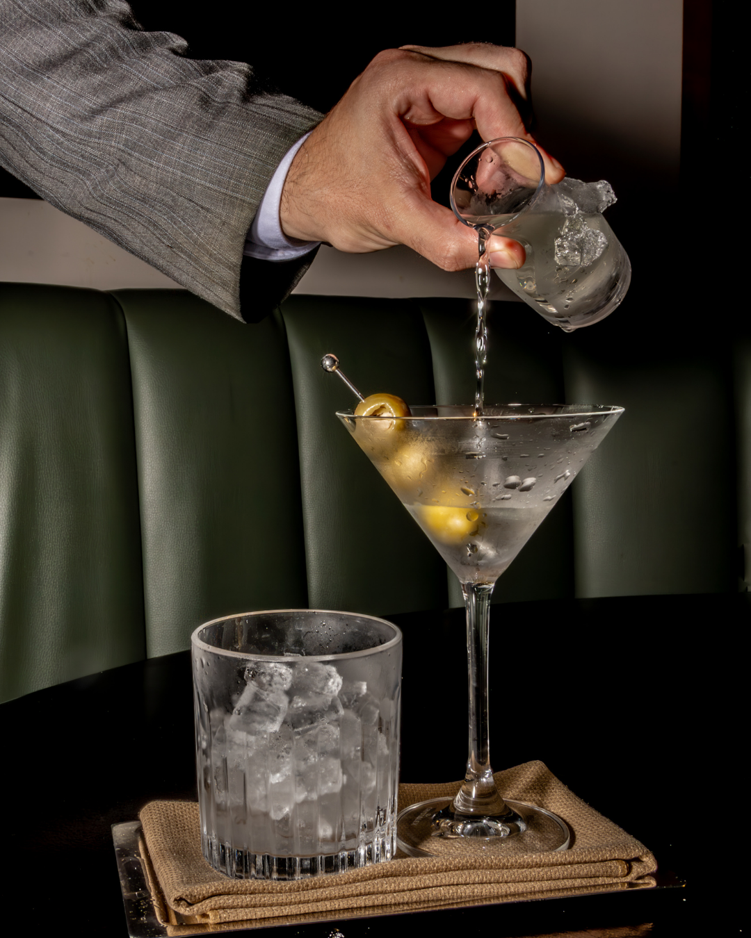 Martini being poured with olives and ice in an elegant bar setting.