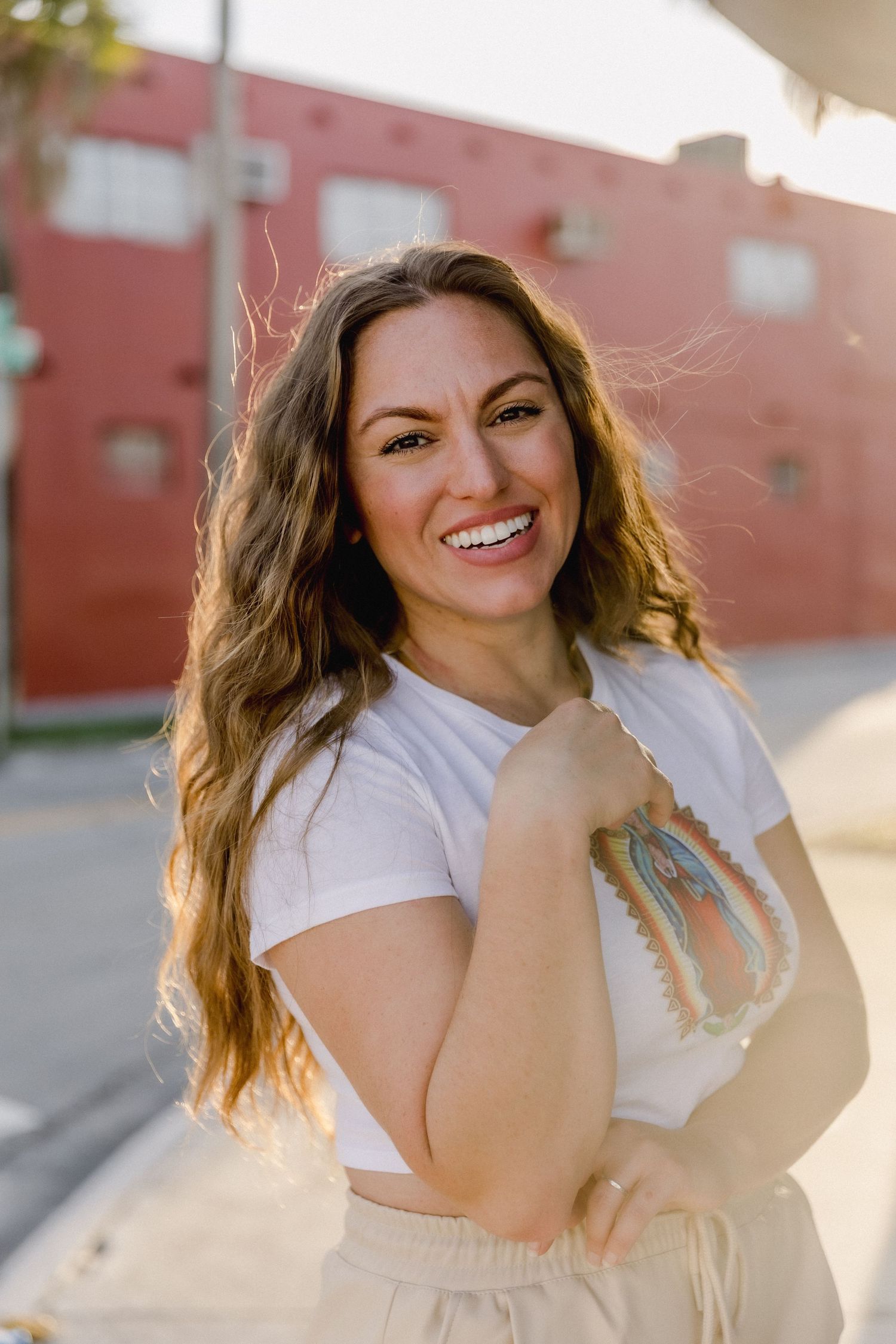 A woman in a white t-shirt smiles and poses outdoors against a brick wall in warm sunlight.