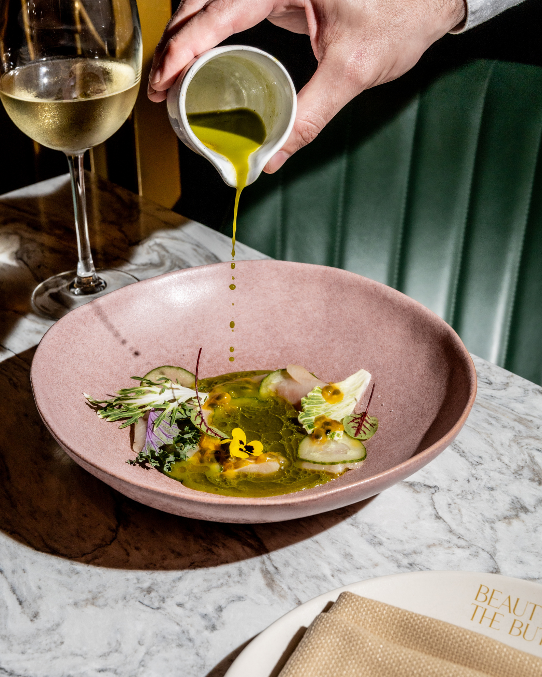 White sauce being drizzled over elegantly plated dishes on pink plates with green banquette seating.