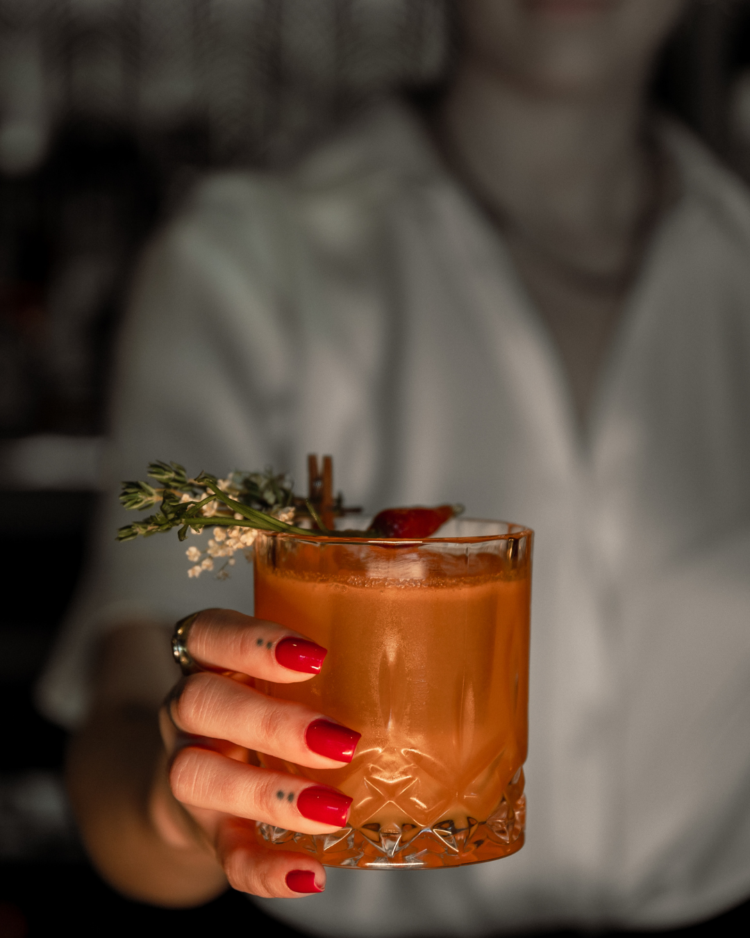 Orange-colored cocktail garnished with fresh herbs and held by manicured hand.