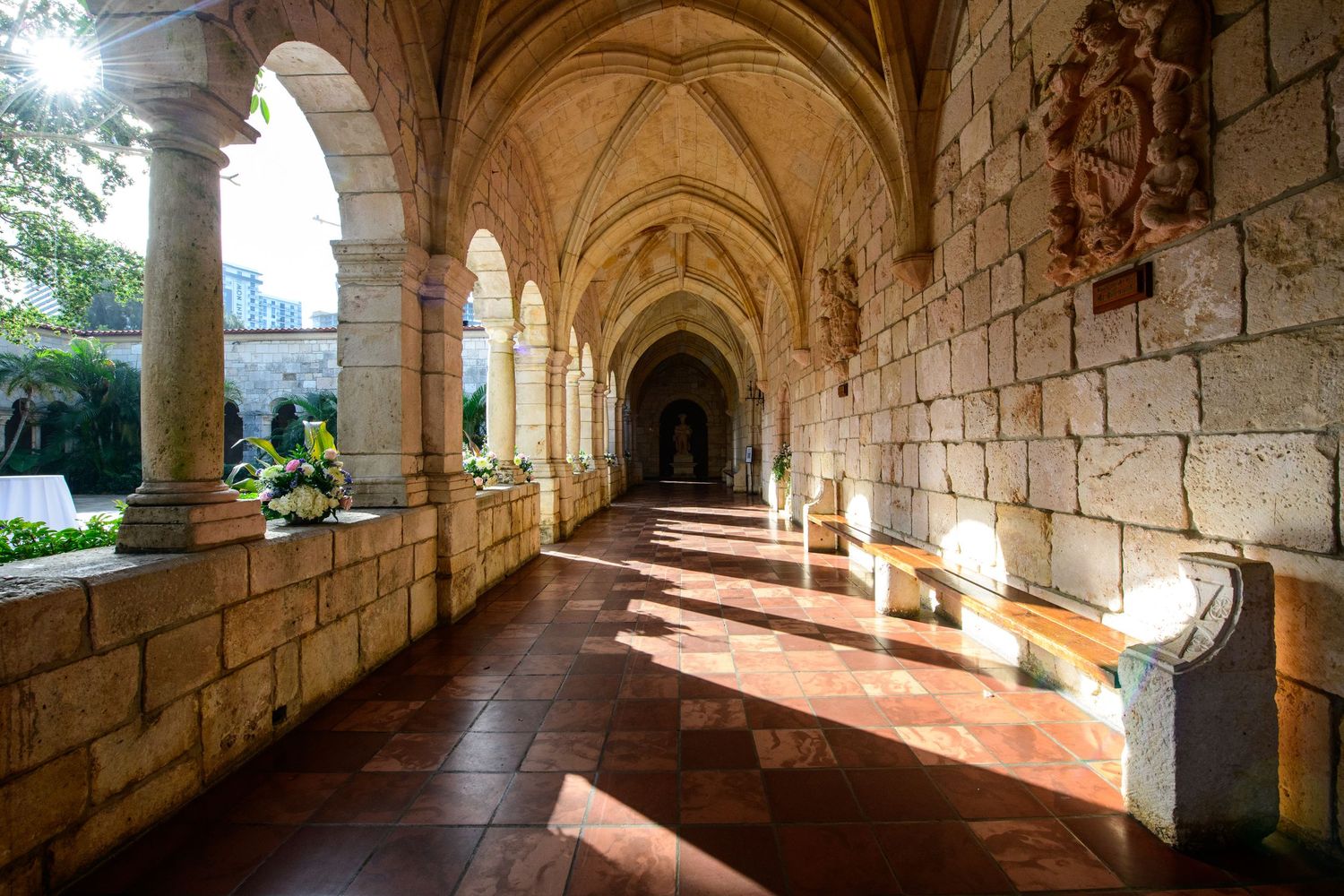 Sunlit medieval stone cloister with vaulted archways and stone benches casting dramatic shadows along corridor.