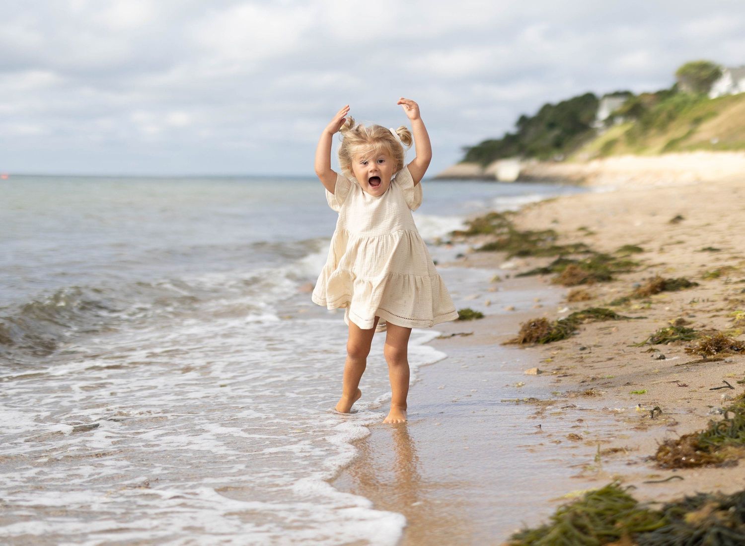 Joyful toddler girl in cream dress celebrating with arms raised on a sandy beach.