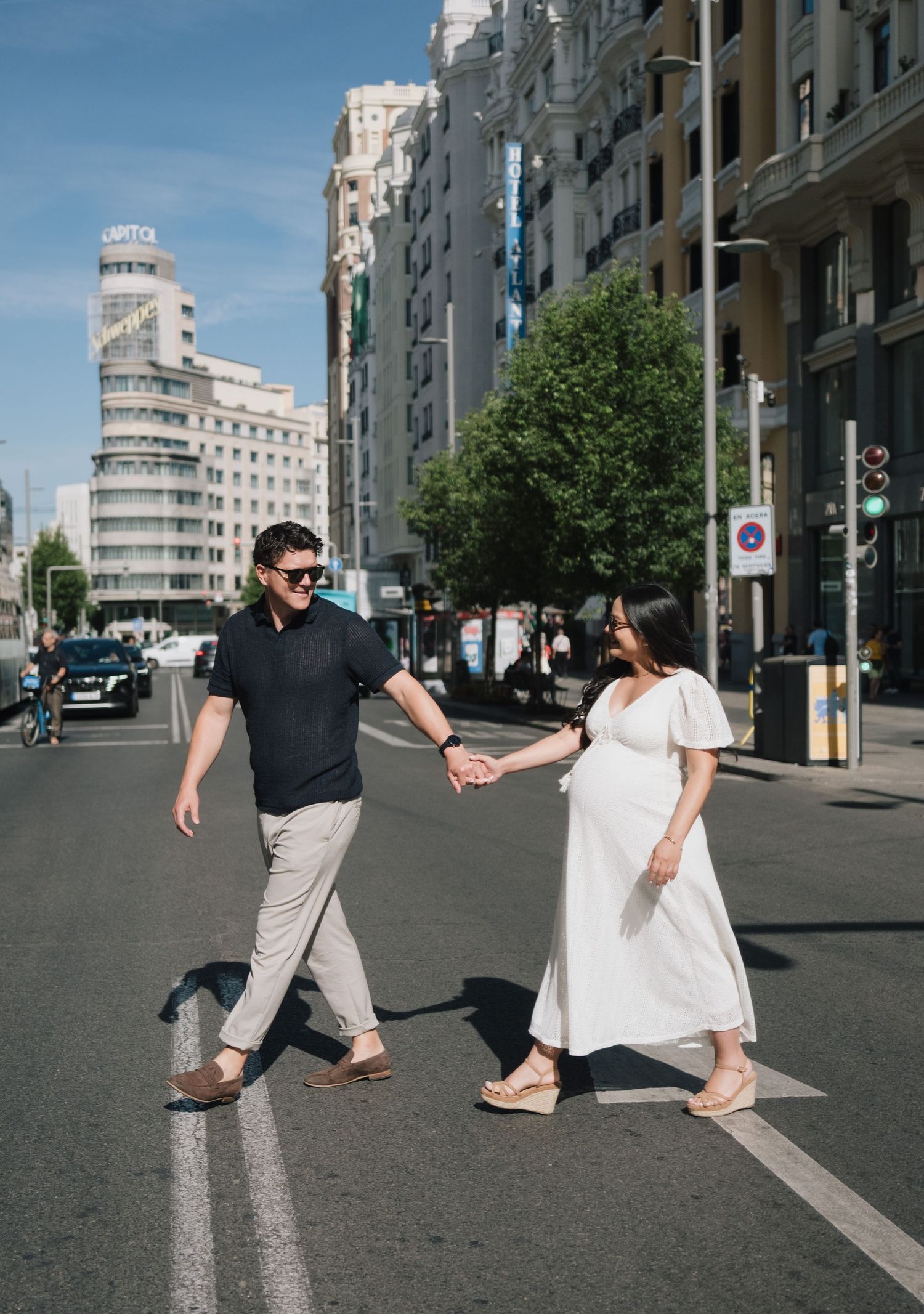 Pregnant couple holding hands crossing a sunny city street in Madrid with urban buildings in the background.