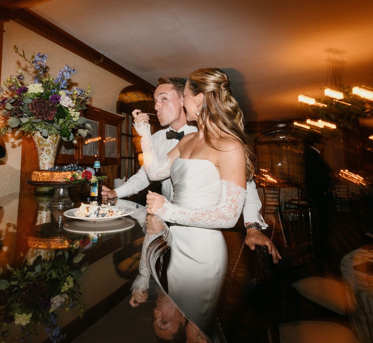 Bride and groom cutting wedding cake together in elegant reception venue with floral arrangements.