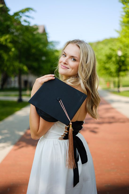 Blonde graduate in white dress holding black cap on sunny brick campus walkway.