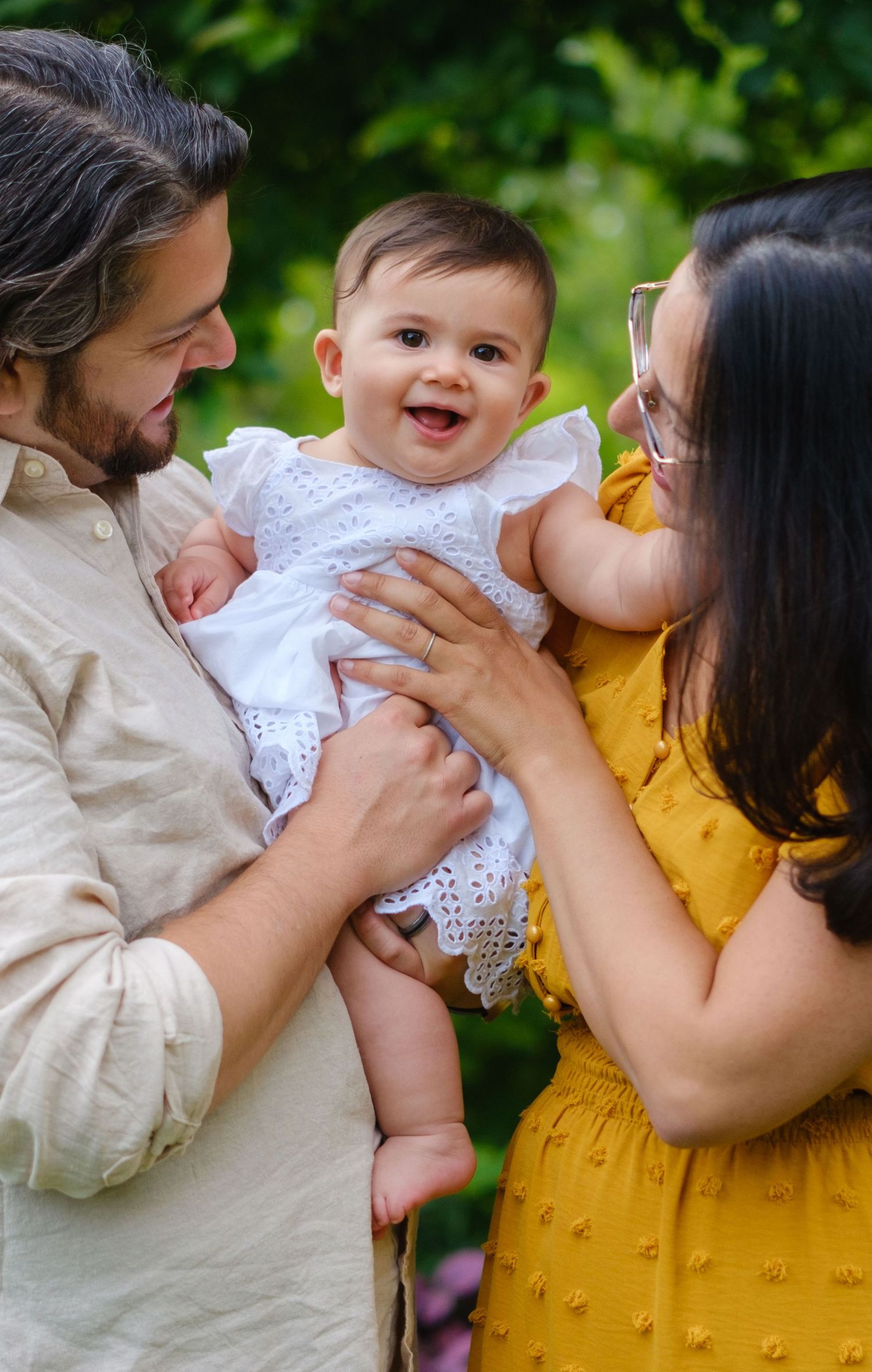 Happy parents holding their smiling baby girl in a white dress outdoors in a lush green garden.