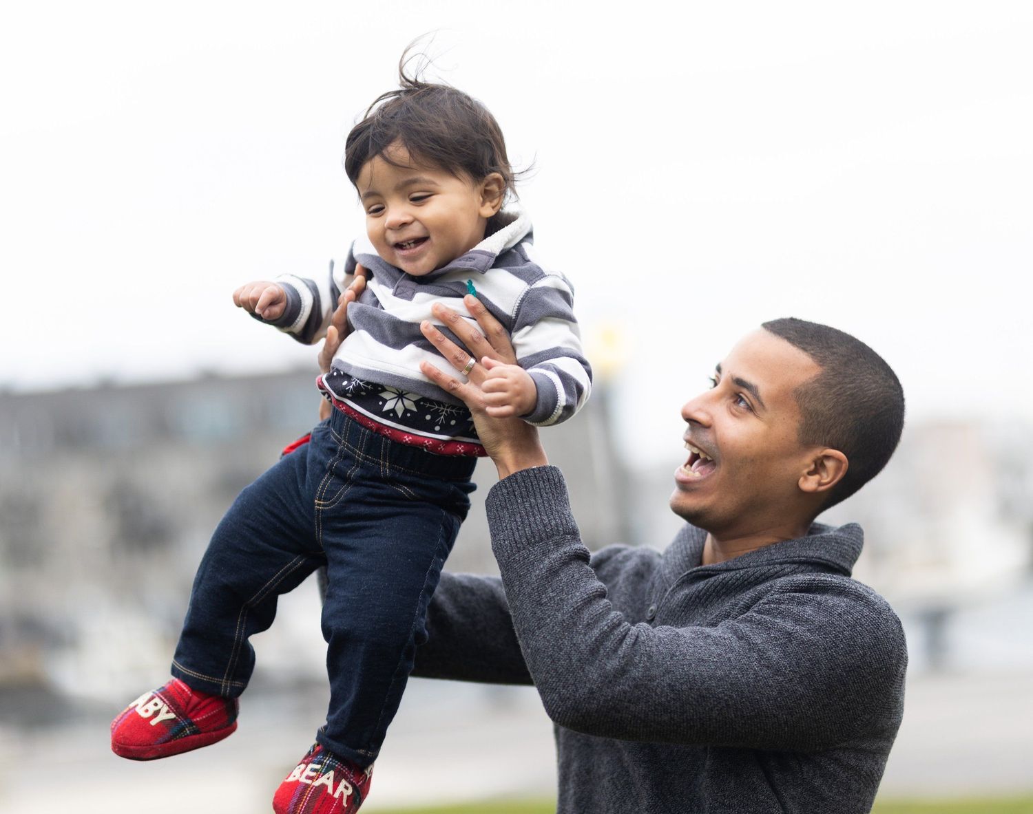 Laughing father lifting happy toddler in striped shirt outdoors on overcast day.