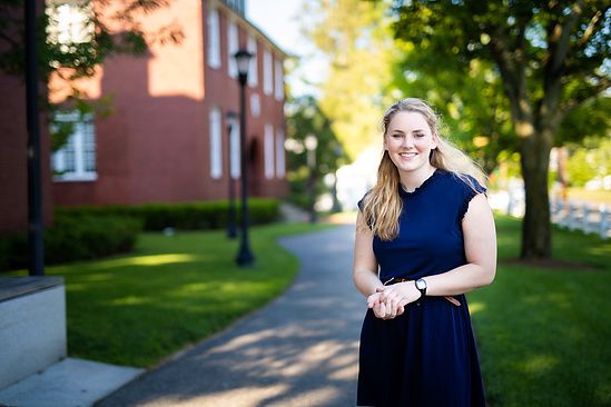 Smiling blonde woman in navy dress standing on a tree-lined brick campus pathway.