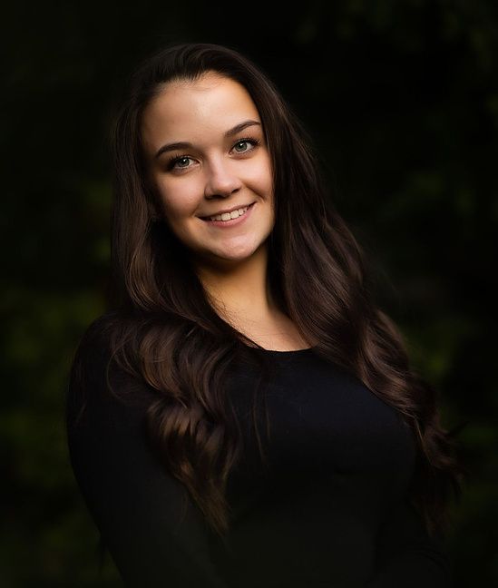 Beautiful brunette woman smiling confidently in black top against lush green background.