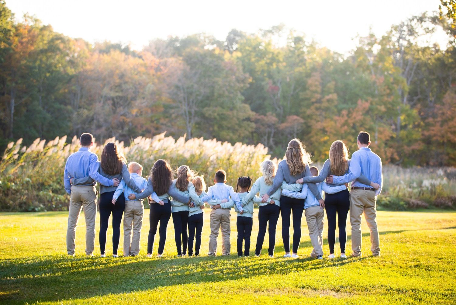 Large family of ten holding hands in a row from behind in a scenic autumn meadow at golden hour.