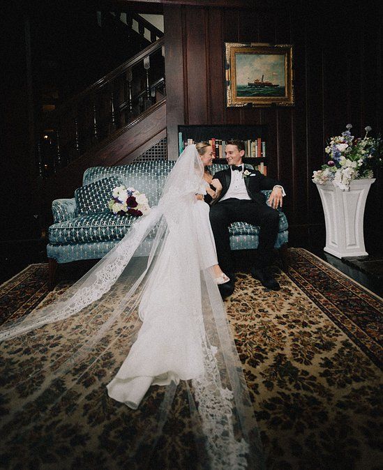 Bride in lace veil and groom in tuxedo share romantic moment on ornate vintage sofa.