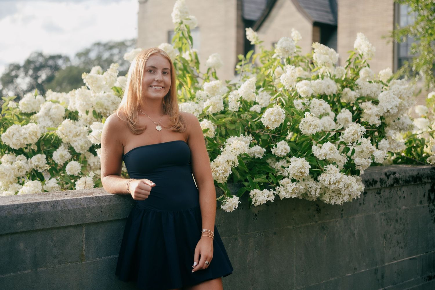 Smiling blonde woman in black dress posing beside white hydrangea blooms outdoors.