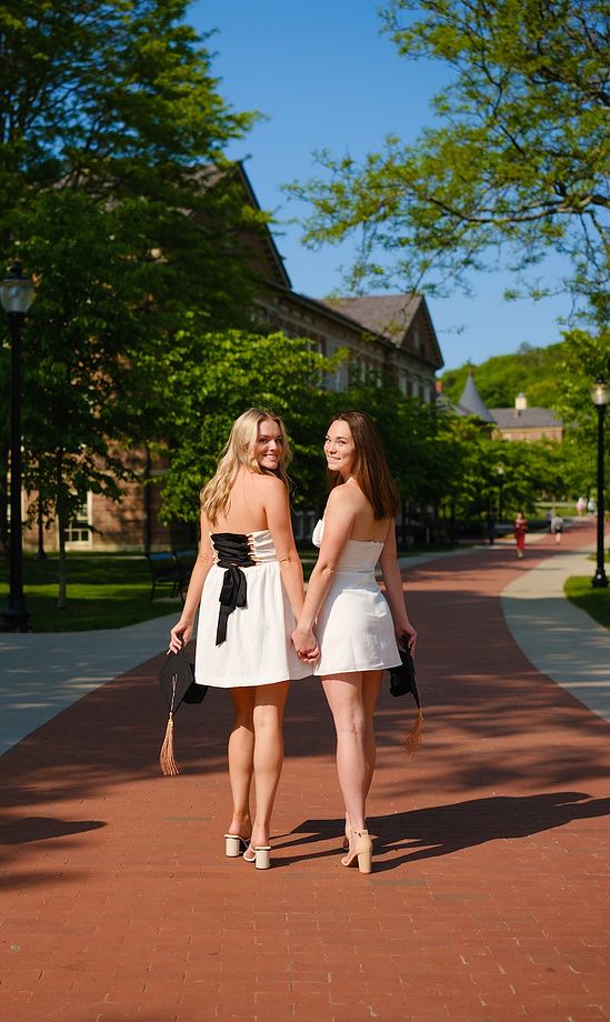 Two smiling women in white dresses hold hands on a brick campus walkway on a sunny day.