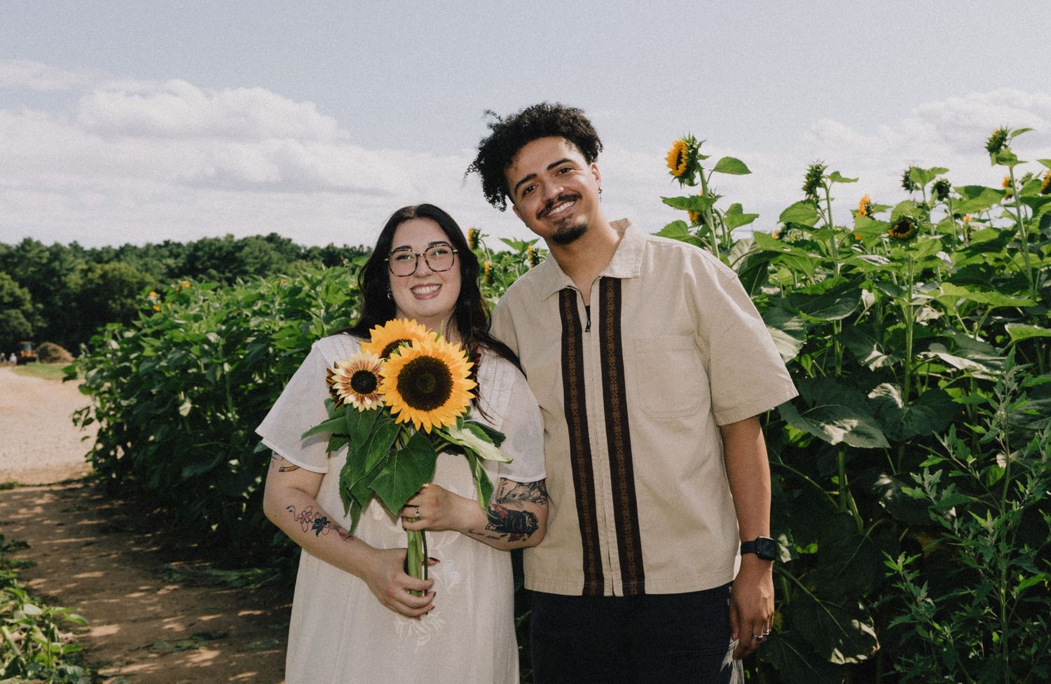 Smiling couple poses together holding sunflowers in a lush green sunflower field.