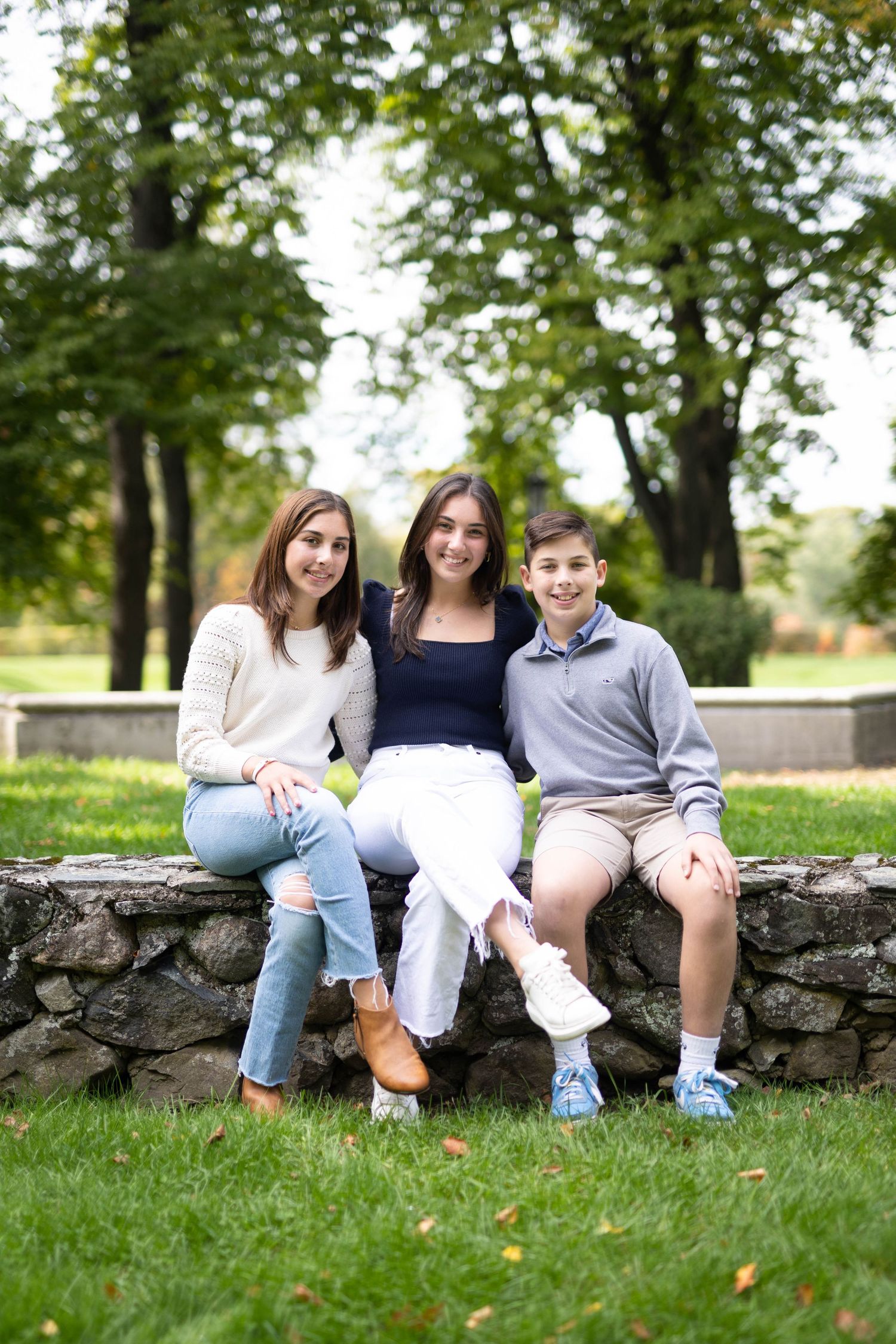 Three teenage siblings sitting on a stone wall smiling together in a lush green park setting.
