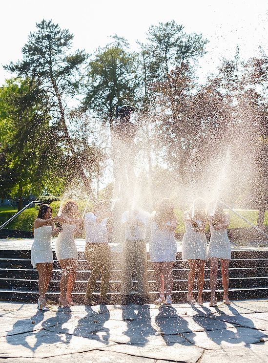 Group of women in white dresses celebrating with confetti spray outdoors on sunny steps.