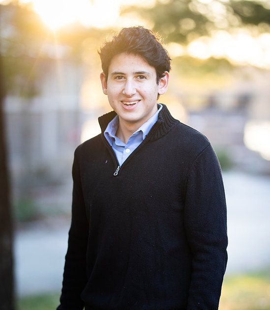 Smiling young man in black zip sweater outdoors with golden sunset bokeh background.