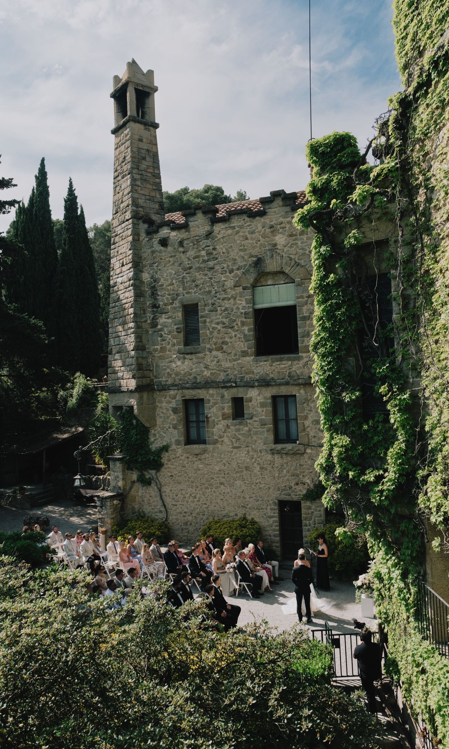 Wedding ceremony in courtyard of historic ivy-covered stone castle with bell tower and cypress trees.