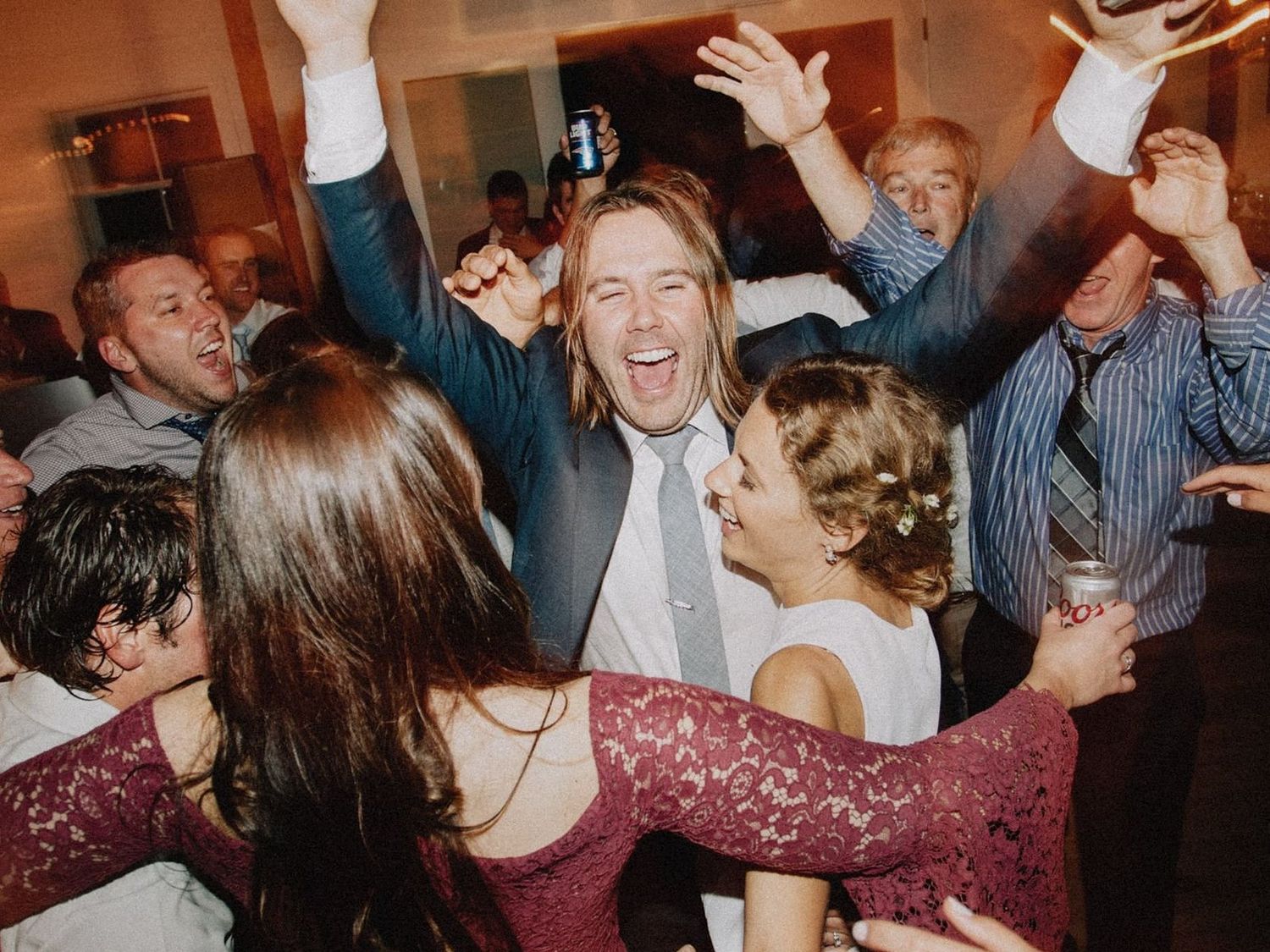 Energetic wedding guests dance and celebrate with arms raised on a crowded reception dance floor.