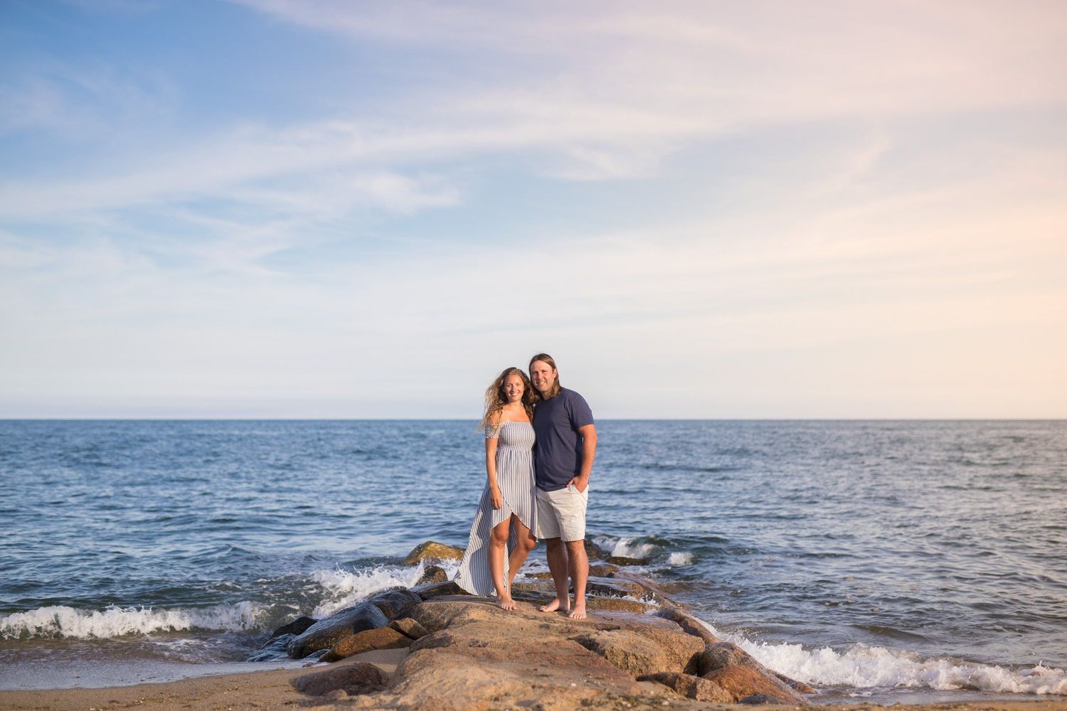 Couple standing together on rocky shoreline with calm ocean and pastel sky behind them.