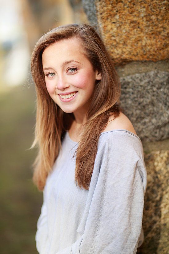 Smiling teenage girl with long brown hair leaning against stone wall outdoors.