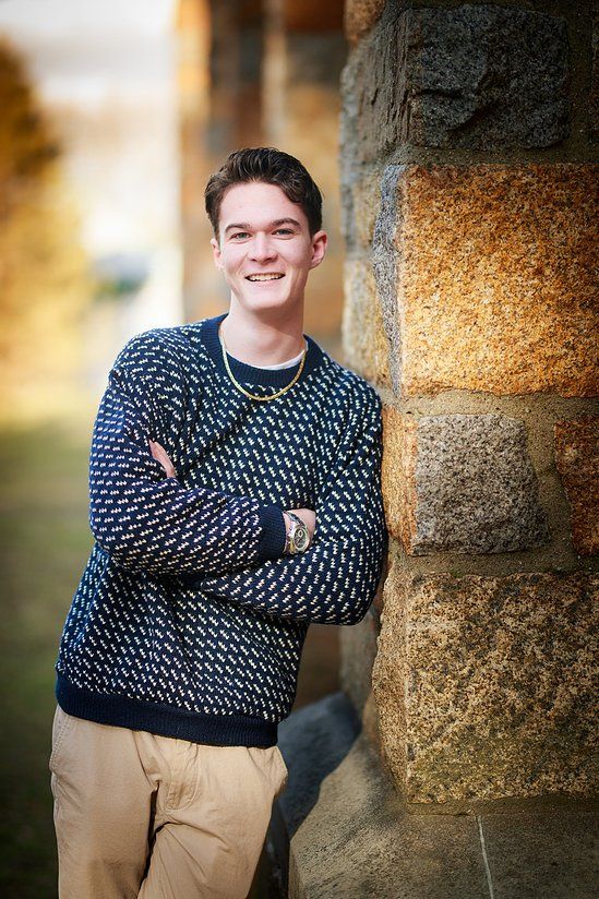 Smiling young man in patterned sweater leaning against stone column with arms crossed.