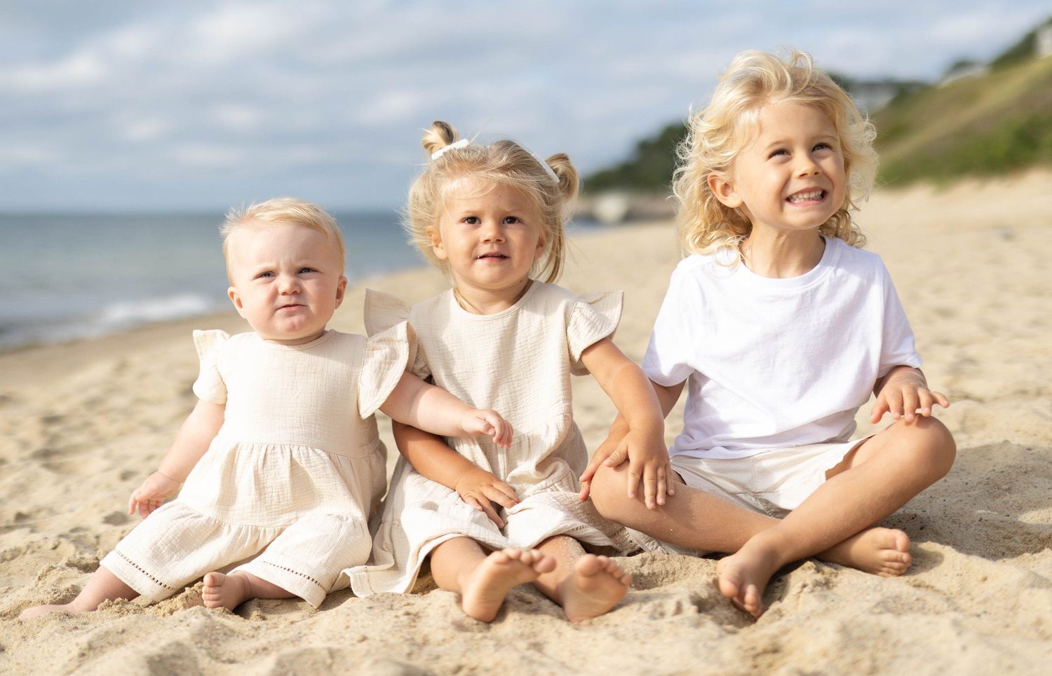 Three young toddler girls sitting together on a sandy beach smiling in white and cream outfits on a sunny day.